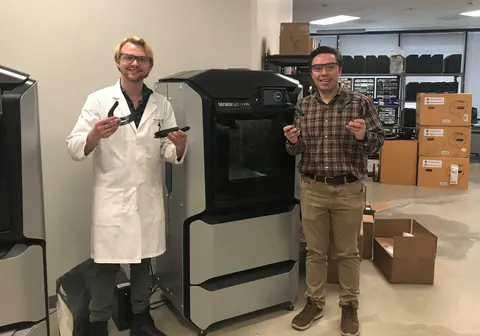 Two Neuralog team members holding 3D-printed face shield parts next to Stratasys 3D printers